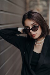 a brunette girl in a black jacket and dress and black glasses walks down the street in the city with a handbag