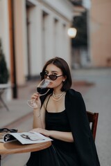 a brunette girl in a black jacket and dress and black glasses sits in a cafe with a handbag and a magazine and drinks wine