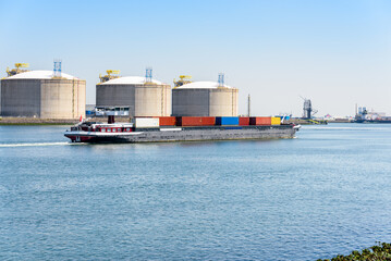 Container barge with harbourside lquefied natural gas tanks in background on a clear summer day. Port of Rotterdam Port, Netherlands.