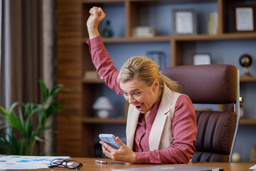 Happy businesswoman surprised by the good news and making yes gesture while looking at smartphone sitting at office. Manager enjoying success, celebrating victory,make good deal, get profit