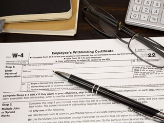 An IRS W-4 Employees Withholding Certificate form, issued in 2022, is shown on display up close, among accounting books, a calculator, glasses, and an ink pen.