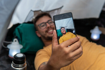 Young man inside his tent in a forest taking a selfie lying down in it