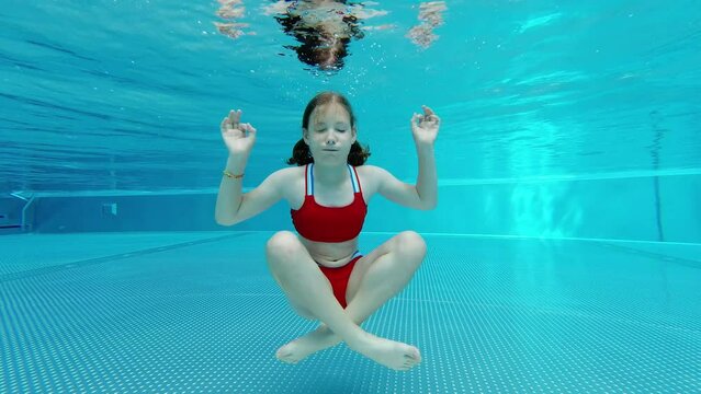 4K slow motion video of teen girl in red swimsuit sitting under water in lotus yoga pose in swimming pool