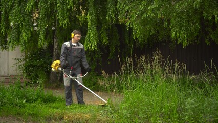 A young man from the special services mows the lawn with a petrol trimmer. A man in glasses and headphones with a lawn mower in his hands.