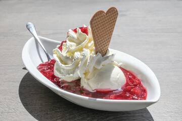 Ice cream with whipped cream, wafer cookie and red fruit jelly, in Germany called rote gruetze, served in a white bowl on a gray table on a sunny summer day, copy space