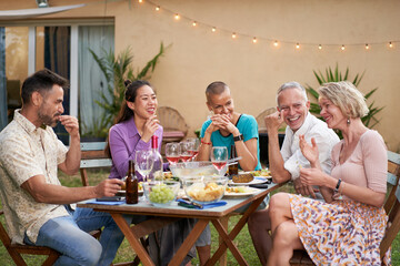 Group of happy friends having fun drinking red wine at barbecue picnic garden party. Middle aged people eating a tasty meal in the backyard.