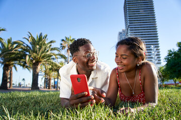 A cheerful young African American couple using a mobile smart phone lying on the grass in the city park. Smiling boy and girl holding cell. Black people using digital devices outdoors.