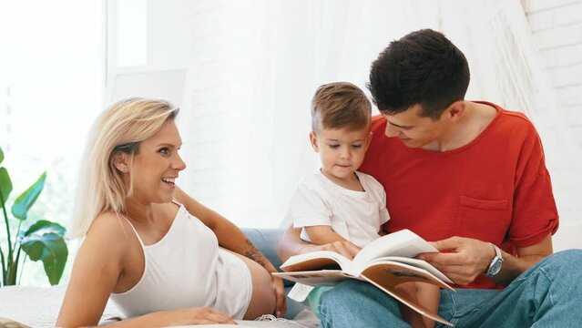 happy family - pregnant mother, father and little son reading a book at home, family concept. High quality 4k footage