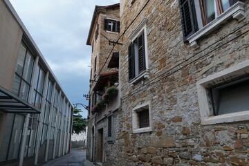 View on a narrow street with old stone building and new modern house in center of Koper, Slovenia. Between buildings is blue sky with white clouds. Old house has a balcony with flowers.