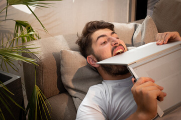 image fun et amusante d'un jeune homme allongé sur son canapé qui s'énerve à cause de son ordinateur portable. Il devient fou et il mord le clavier