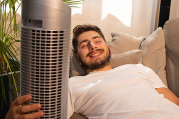 jeune homme allongé sur son canapé. Il est heureux car il profite de son ventilateur pendant la...