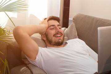 Jeune homme souriant allongé dans son canapé qui utilise un ordinateur portable pour travailler,...