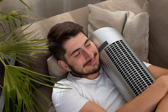 image fun et amusante d'un jeune homme qui embrasse son ventilateur pendant la canicule. Il est allong&eacute; dans son canap&eacute; et il fait chaud.