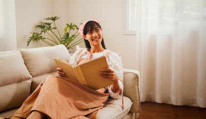 Portrait young asian woman sits happily reading large novel on the sofa in her home favorite story...