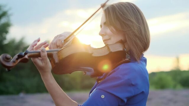 Close-up adult caucasian woman violinist playing violin on summer lavender field in evening, romantic musician in blue dress enjoying walking on nature