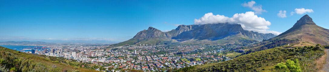 Fototapeta na wymiar Panoramic landscape of Table mountain and surrounding urban town and a scenic road for traveling along Cape Town, South Africa. A mountain road overlooking the city with a cloudy blue sky in summer