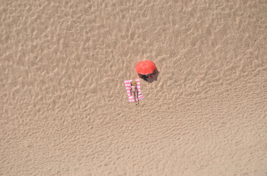Rapariga deitada na praia com guarda sol ao lado aberto e chap&eacute;u de palha a tapar a cabe&ccedil;a, vista a&eacute;rea