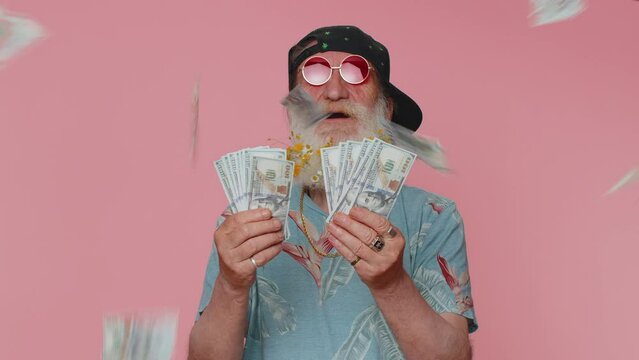 Rich pleased boss senior man waving money dollar cash banknotes bills like a fan, success business career, lottery winner, big income, wealth. Elderly grandfather isolated on pink studio background