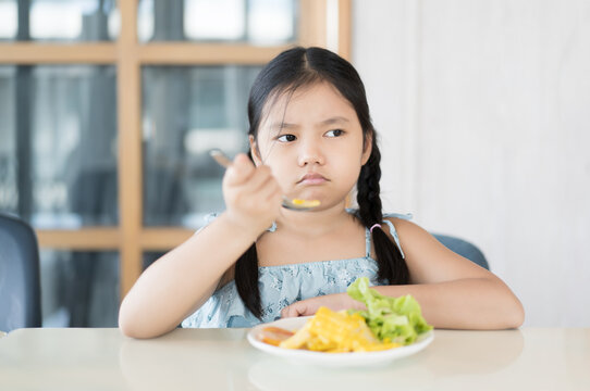 Asian child frowning or kid girl unhappy and don't like eating vegetables salad with corn tomato and greens vegetable with holding spoon or person anorexic bored food at kitchen school or restaurant