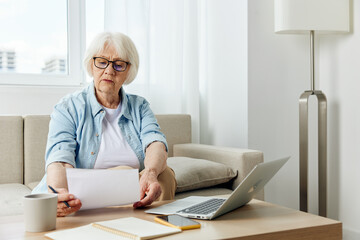 an elderly lady works sitting on the couch at home reading a text from a paper during an online conference with colleagues