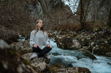 Girl sits on a large stone and meditates near a waterfall next to a mountain river.