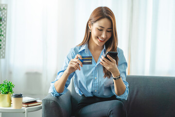 Young smiling Asian woman using smartphone buying online shopping by credit card while lying on...