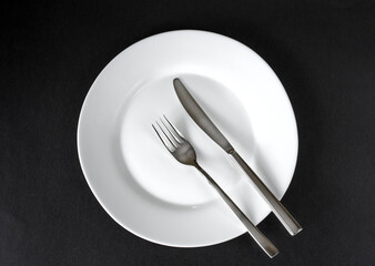 Served plate with cutlery. Empty white plate and stainless knife and fork isolated on black background. Top view.