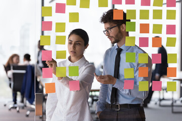 Diverse millennial employees, coworkers discussing project schedule, writing memo on sticky notes....