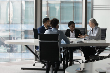 Fototapeta na wymiar Mature business teacher, mentor training diverse team of interns, talking to group of students at meeting table in modern office space. Millennial business colleagues discussing at glass wall window