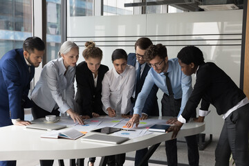 Diverse different aged business group standing at meeting table, negotiating on marketing reports,...