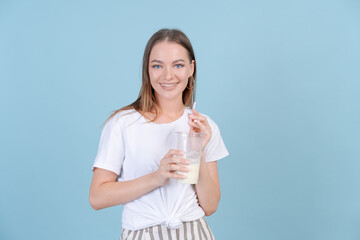 Cute caucasian young girl in white t-shirt and striped pants drinks milkshake on blue background and smiles with her teeth while looking at the camera