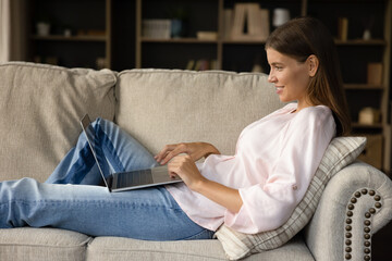 Side view woman sits on couch in living room with laptop enjoy on-line chat, share text messages in social media, communicates remotely use internet. Modern tech, fun, leisure, telecommuting concept