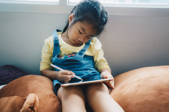 Asian Kid Girl Drawing With Tablet Computer In Cozy House