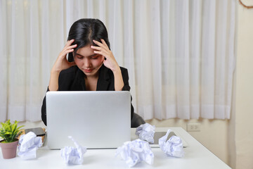 Mid adult businesswoman touching head on table after bad news business failure or get fired and feeling discouraged, distraught and hopeless on blue background with computer and crumpled paper