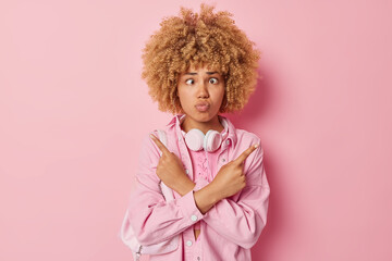 Hesitant confused young woman makes funny grimace tries to make choice crosses hands against chest points left and right dressed casually isolated over pink background. What to choose I wonder