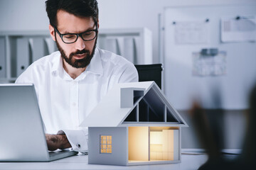 Businessman typing on laptop looking at model of paper house