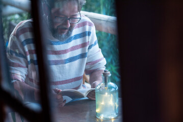 View of a man reading a book through the glass window from inside home. Concept of people and education or relax leisure reading activity. One person studying outdoor on the terrace