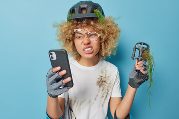 Irritated woman with curly hair clenches teeth looks annoying at smartphone holds pedal tries to find in internet how to repair bicycle stands dirty after accident isolated over blue background