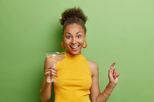 Positive Carefree Beautiful Woman With Curly Dark Hair Dances With Glass Of Cocktail Wears Yellow T Shirt And Earrings Has Positive Mood Isolated Over Vivid Green Background Enjoys Party Time