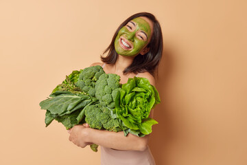 Positive young Asian woman with dark hair embraces heap of fresh green vegetables applies...