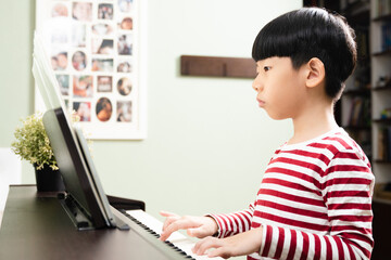 Cute Asian elementary school child learning online with computer tablet, practicing and playing electric piano with concentration at home. Child brain development, music therapy and education.