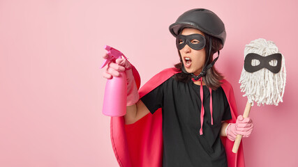 Horizontal shot of angry woman superhero uses spray detergent holds mop ready to save world from dirt exclaims loudly isolated over pink background empty space for your advertisement or promo