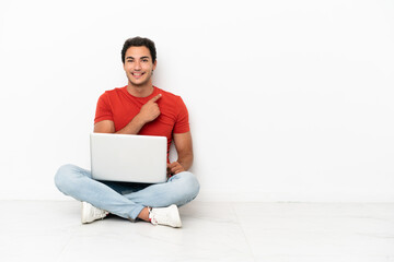Caucasian handsome man with a laptop sitting on the floor pointing to the side to present a product