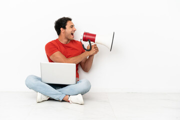 Caucasian handsome man with a laptop sitting on the floor shouting through a megaphone