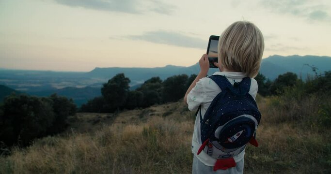Preschool children using smartphone on hike travel adventure. Child boy stand on top of mountain and shoot a picture with mobile phone. Kids using technology on journey