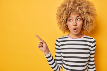 Horizontal shot of shocked curly haired young woman points away on blank space for your...