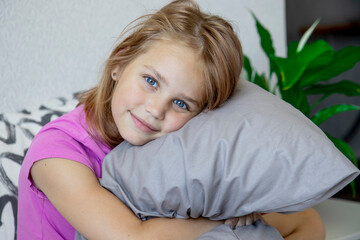 Charming little blonde girl with blue eyes looks into the camera and hugs a pillow while sitting on the bed after sleeping