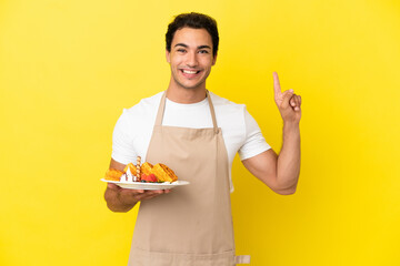 Restaurant waiter holding waffles over isolated yellow background pointing up a great idea