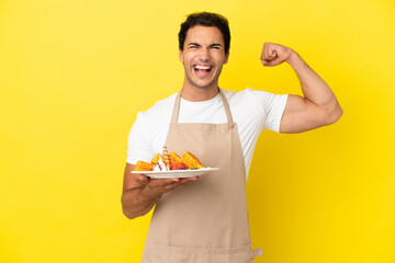 Restaurant waiter holding waffles over isolated yellow background doing strong gesture