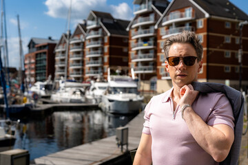 A handsome businessman in a casual grey suit in front of a busy marina with luxury yachts moored up